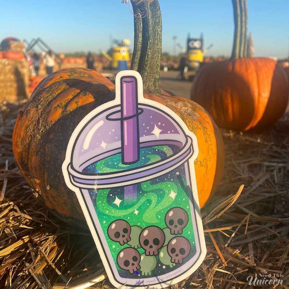Witch’s Brew Boba Sticker photographed at a pumpkin patch. The sticker features glowing green holographic swirls and skull-shaped boba pearls inside a spooky bubble tea cup. Accented holographic finish catches the light. Orange pumpkins and hay bales in the background contrast with the eerie green holographic potion design, enhancing the Halloween aesthetic.